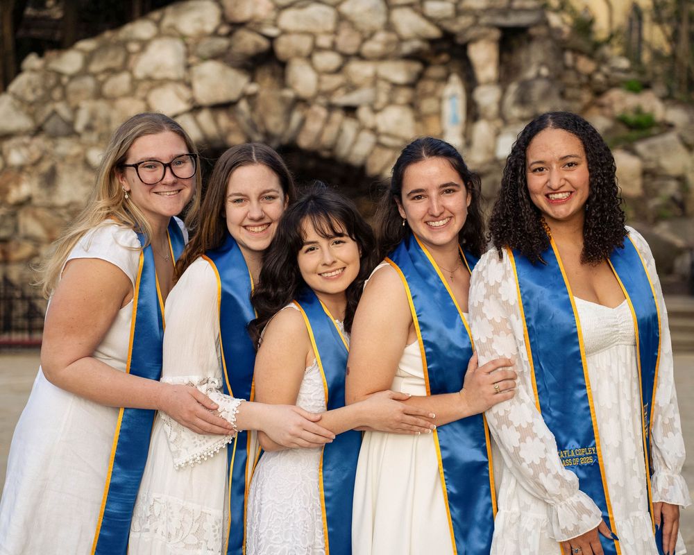 Five graduates in white dresses and blue sashes smile together in front of a stone wall during commencement ceremony.