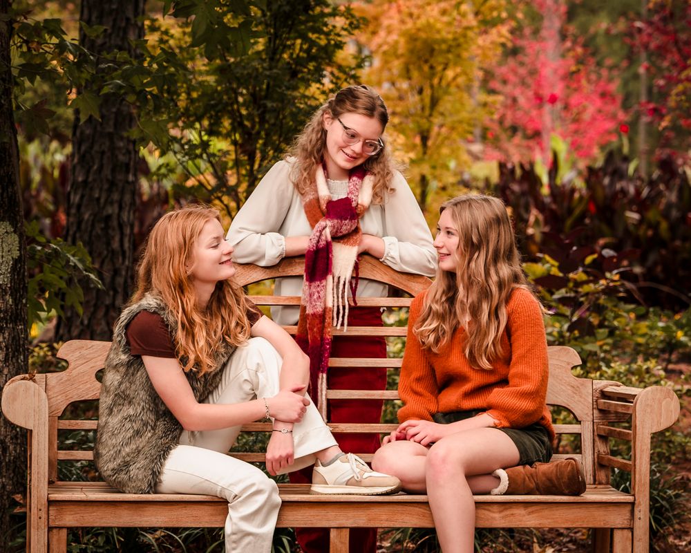 Three people sit on a wooden bench having a conversation in a colorful autumn garden with vibrant foliage.