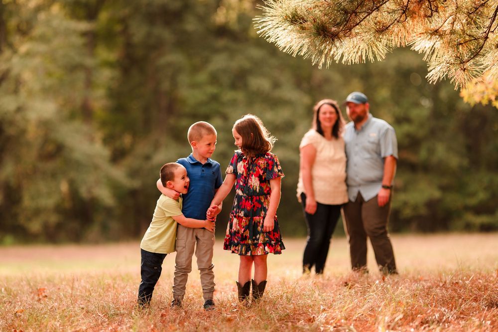 Family portrait outdoors in a golden autumn field at sunset with pine trees in the background.