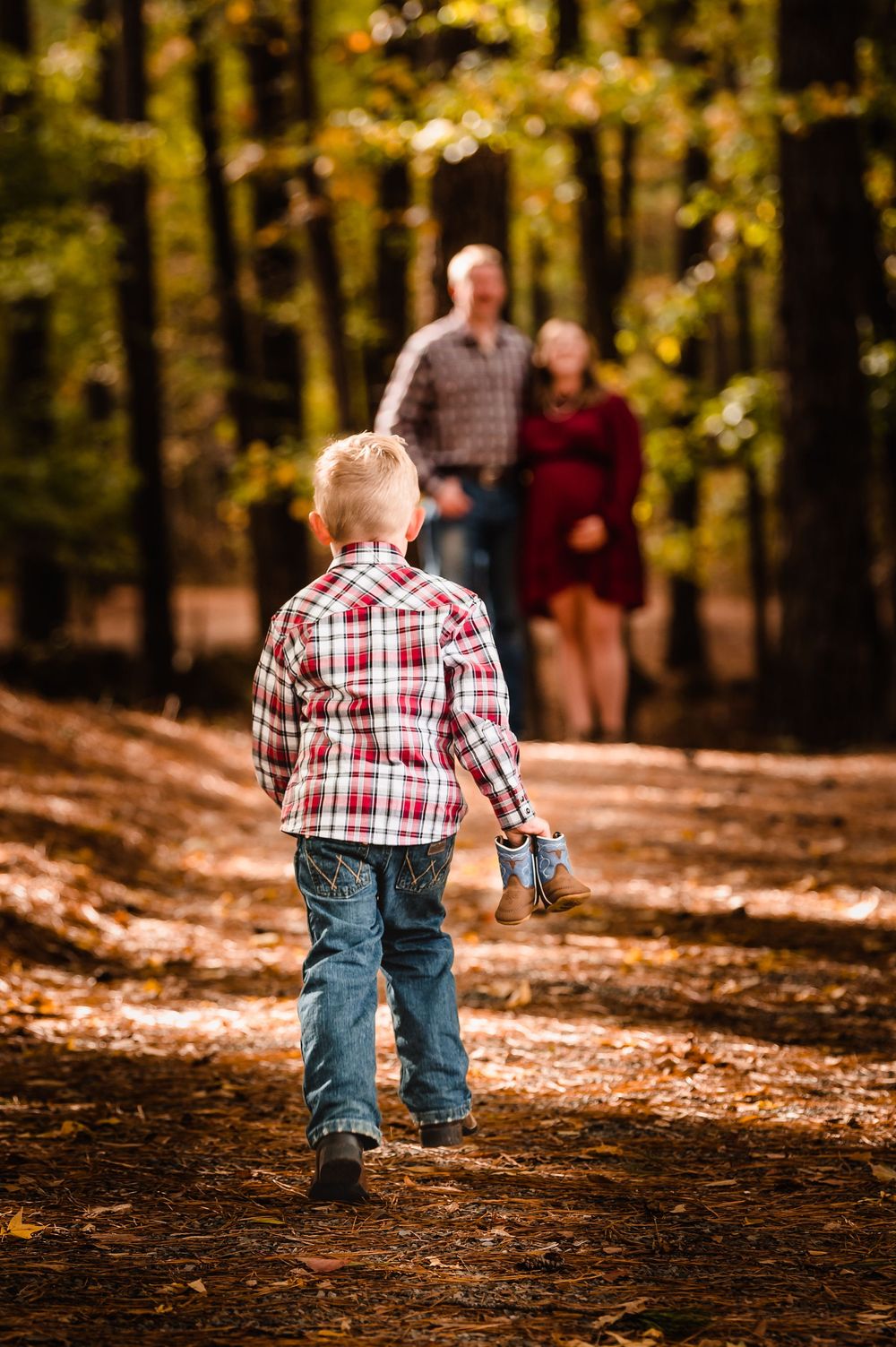 Young child in plaid shirt and jeans walks along autumn forest trail with fallen leaves while two adults follow behind.