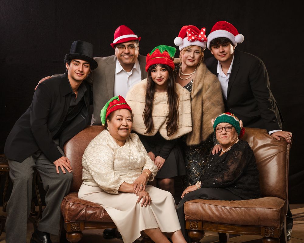 Group wearing festive holiday hats poses together on brown leather couch against dark background.