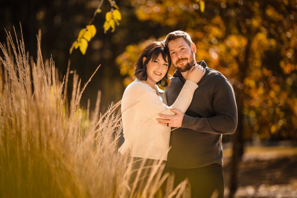 A couple embraces in a golden autumn field during sunset as warm light illuminates the tall grass around them.