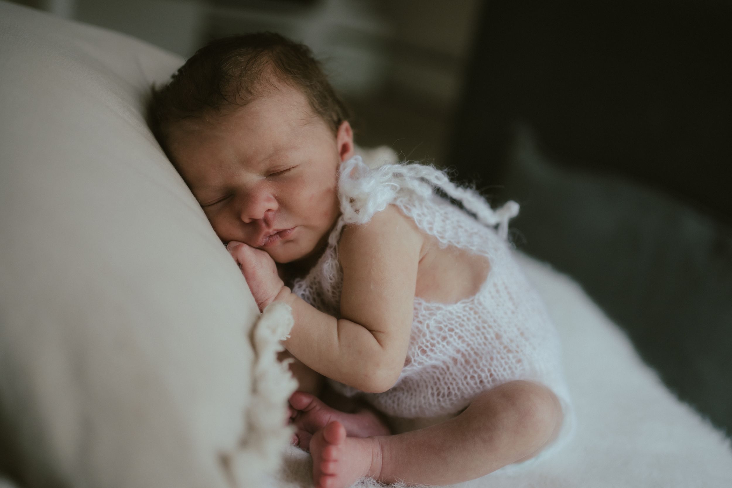 Sleeping newborn baby in white lace outfit rests peacefully on soft cushions.