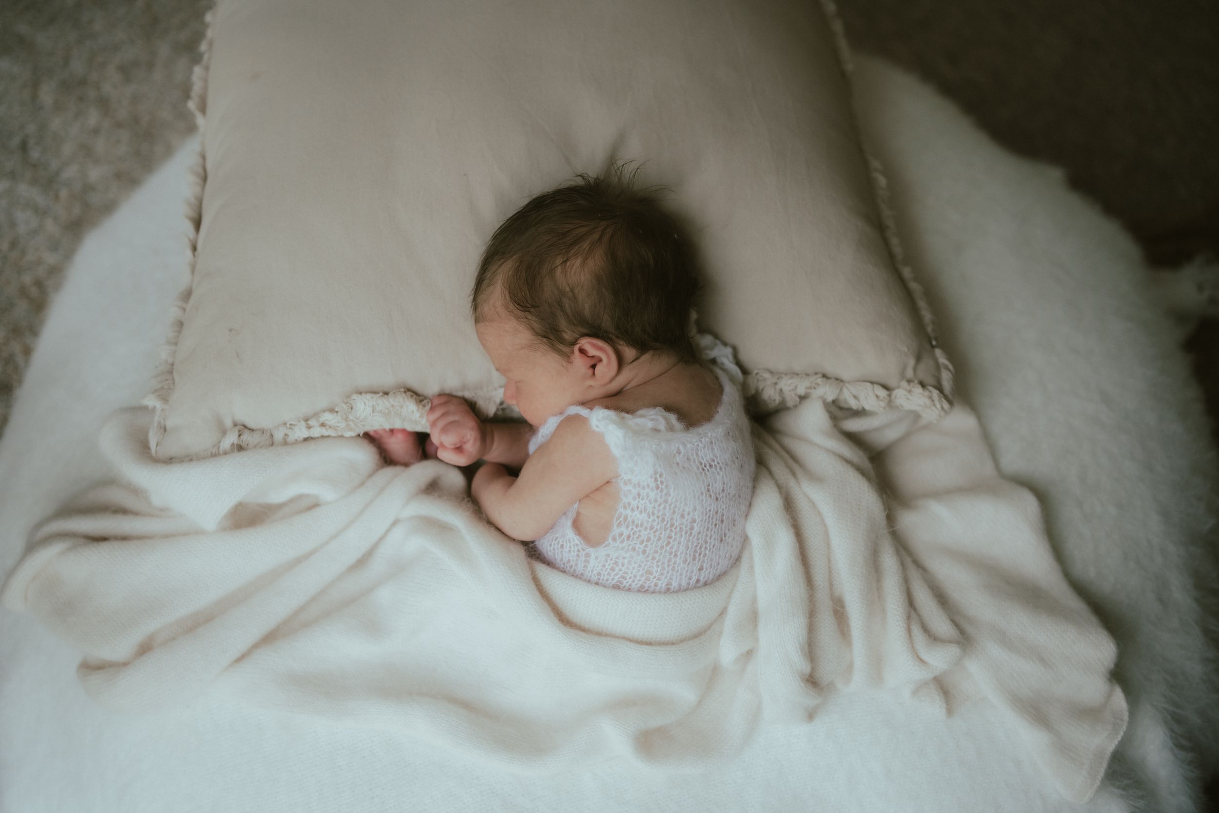 A small baby wearing white sleeps peacefully curled up on soft white bedding.