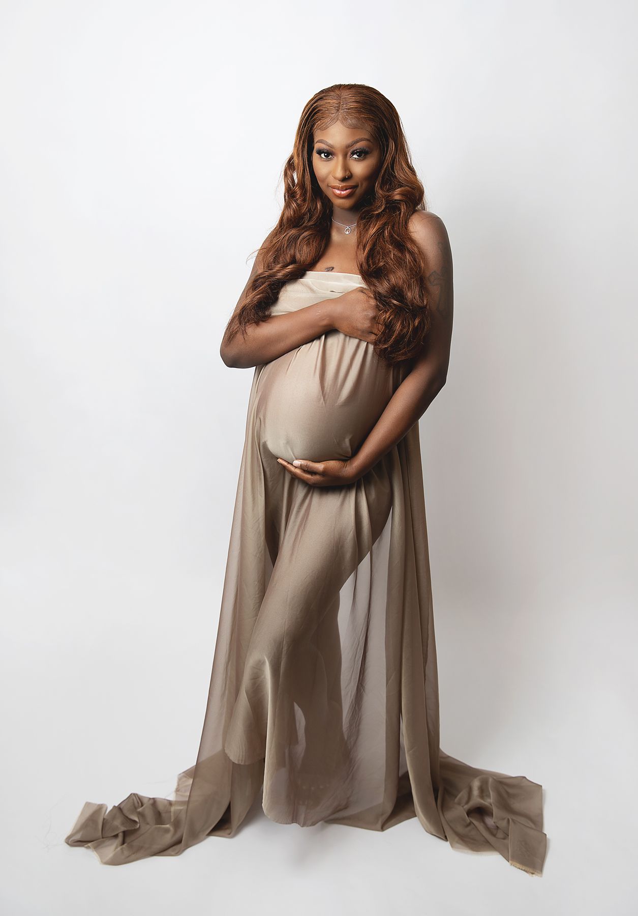 A glowing figure in a flowing taupe dress poses gracefully against a white backdrop for an elegant maternity portrait.