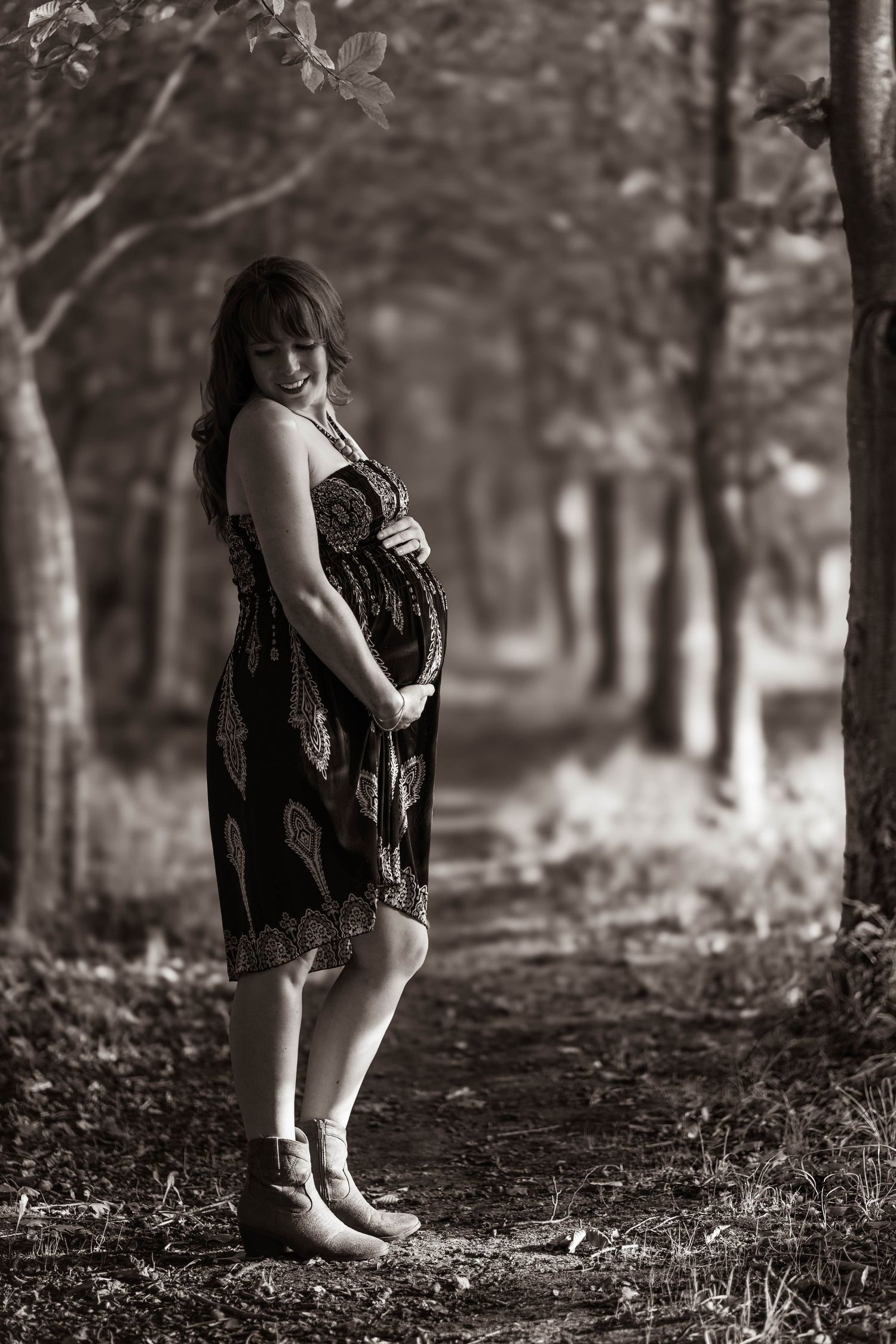 An expectant mother in a patterned dress poses in a serene wooded path.