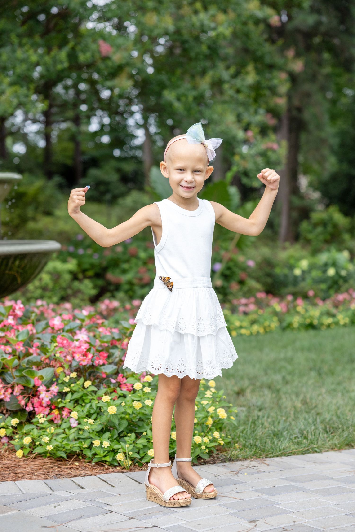 Young child poses with arms raised wearing white summer dress and bow in garden setting with pink flowers. #NOELLESTRONG