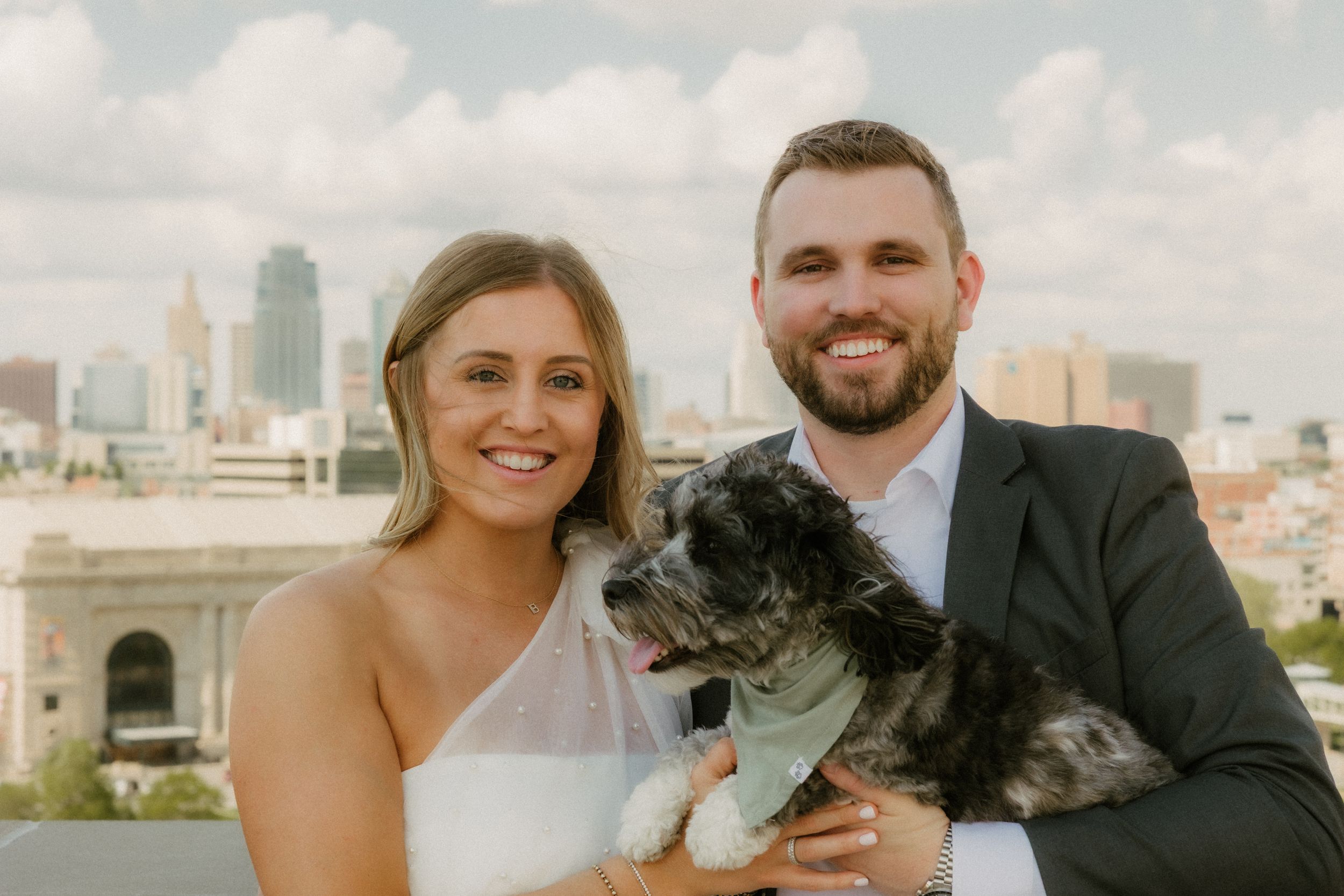 A couple in wedding attire smiles while holding their small black and white dog on a rooftop with a city skyline behind them.