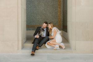 A couple in formal attire sits together against an ornate wall during their wedding photo session.
