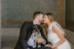 A couple in wedding attire shares a tender moment while holding their small scruffy dog between them.