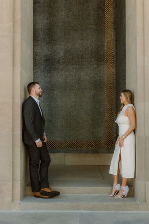 A couple stands facing each other in an elegant archway with a textured grey wall backdrop.