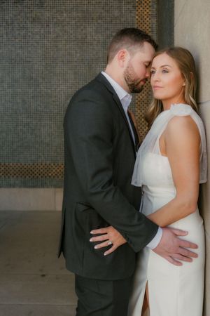 A couple shares an intimate moment against a modern tiled wall in formal wedding attire.