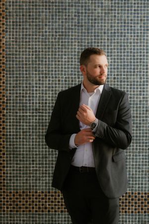 Professional in a dark suit adjusts his shirt cuff while standing against a modern tiled wall background.