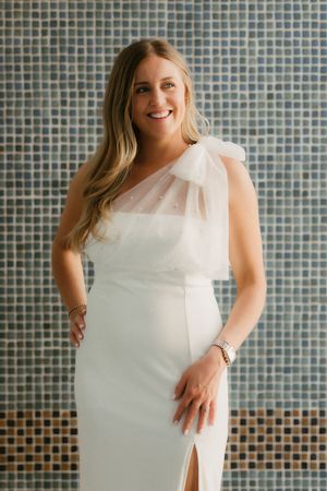 Elegant woman in a white one-shoulder dress with sheer tulle detail poses against a blue mosaic tile wall.