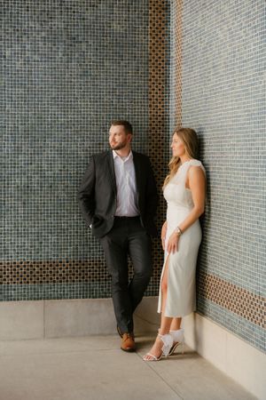 Stylish couple in formal attire pose against modern tiled wall in an urban setting.