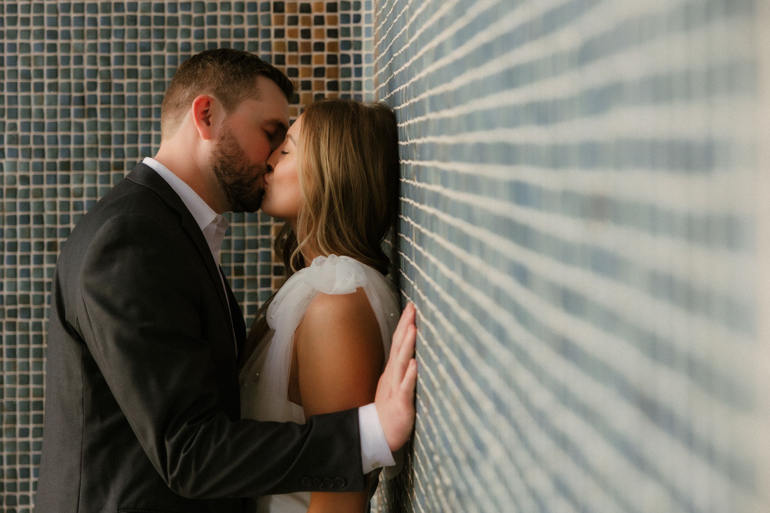 Intimate couple sharing a romantic moment against a tiled wall in formal attire.
