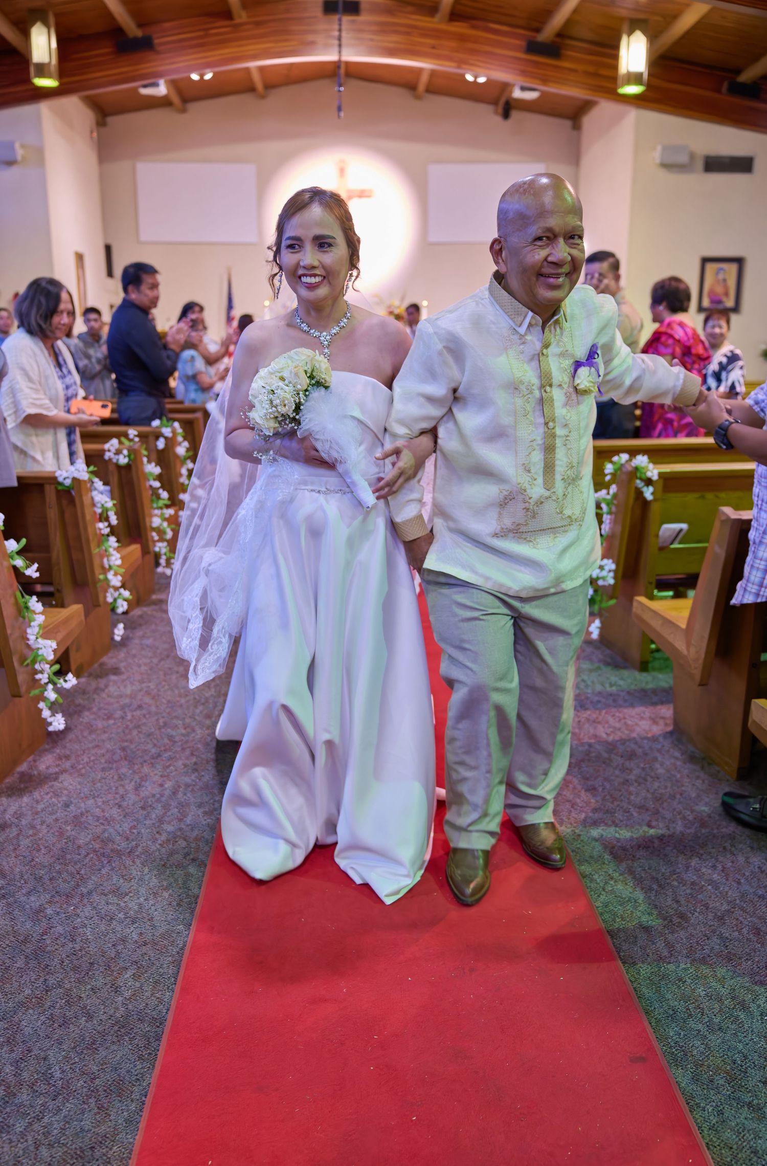 A couple walks down a red carpet aisle after their wedding ceremony in a church with wooden pews and hanging lights.