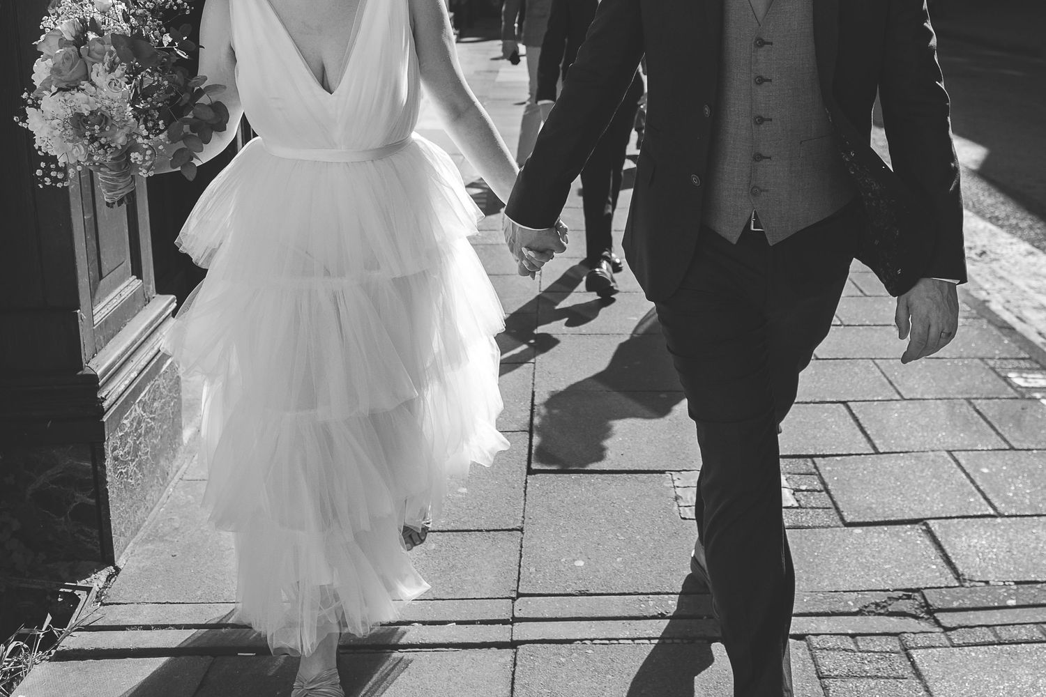 Black and white photo of a bride in a tulle wedding dress walking with groom on a stone pathway in sunlight.