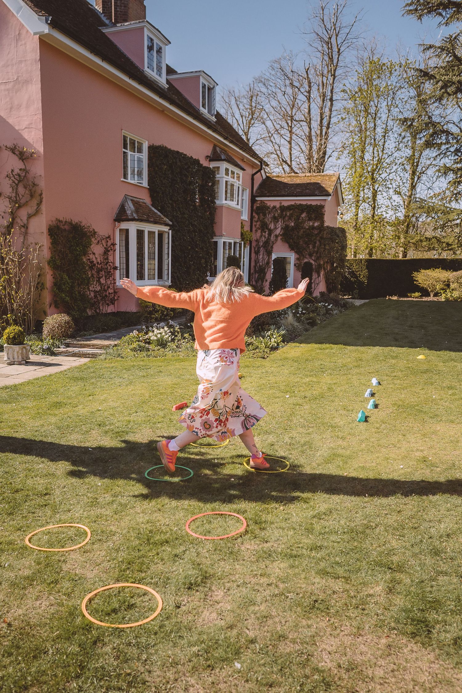 A person jumps playfully through colorful hula hoops laid out on a lawn behind a pink house on a sunny day.