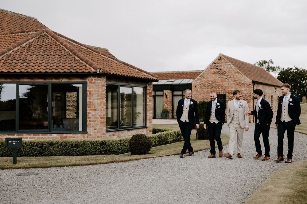 A wedding party in formal attire walks along a path outside a brick building with terracotta roof tiles.