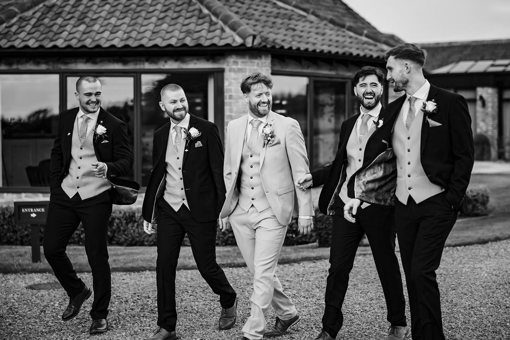 Black and white photo of groom with groomsmen laughing and posing together outside a venue in formal attire.