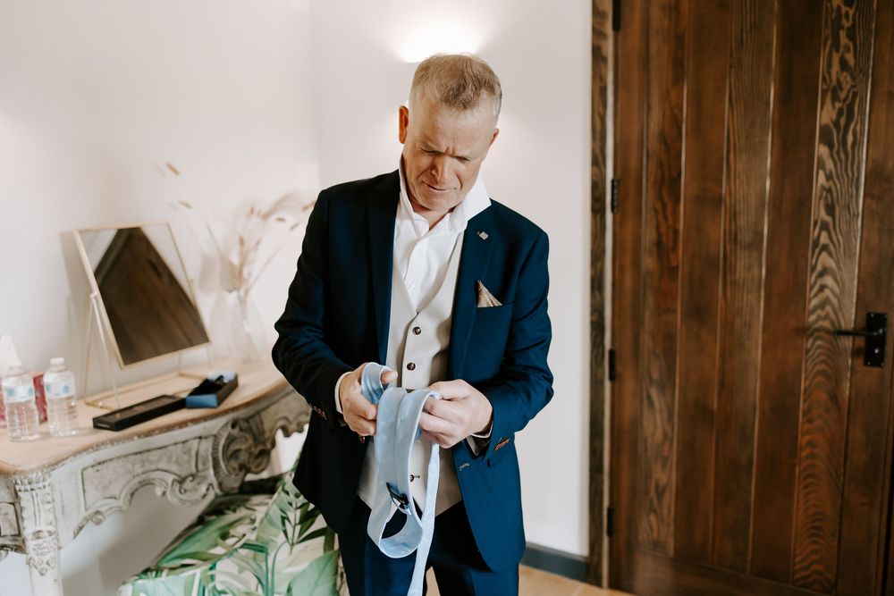 A person in a navy blue suit and white dress shirt holds a lantern while standing in a rustic indoor setting.