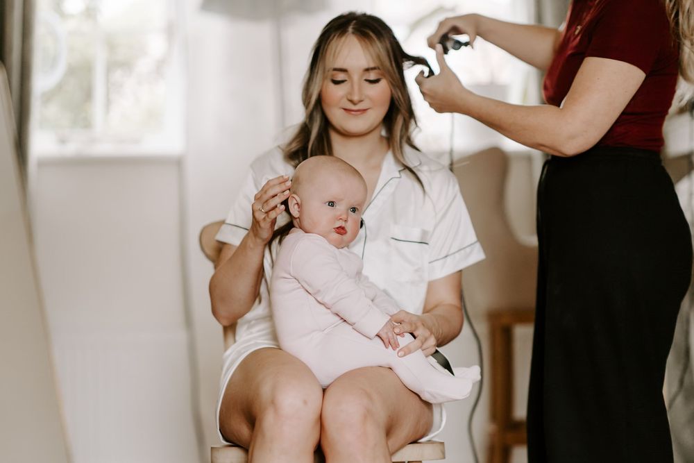 A stylist works on someone's hair during a getting-ready session with an infant present.