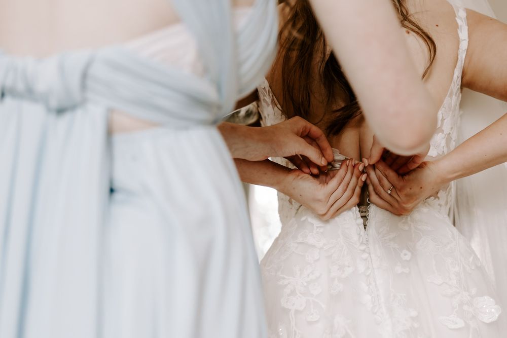 Hands help fasten buttons on a delicate white wedding dress while a light blue bridesmaid dress is visible in frame.