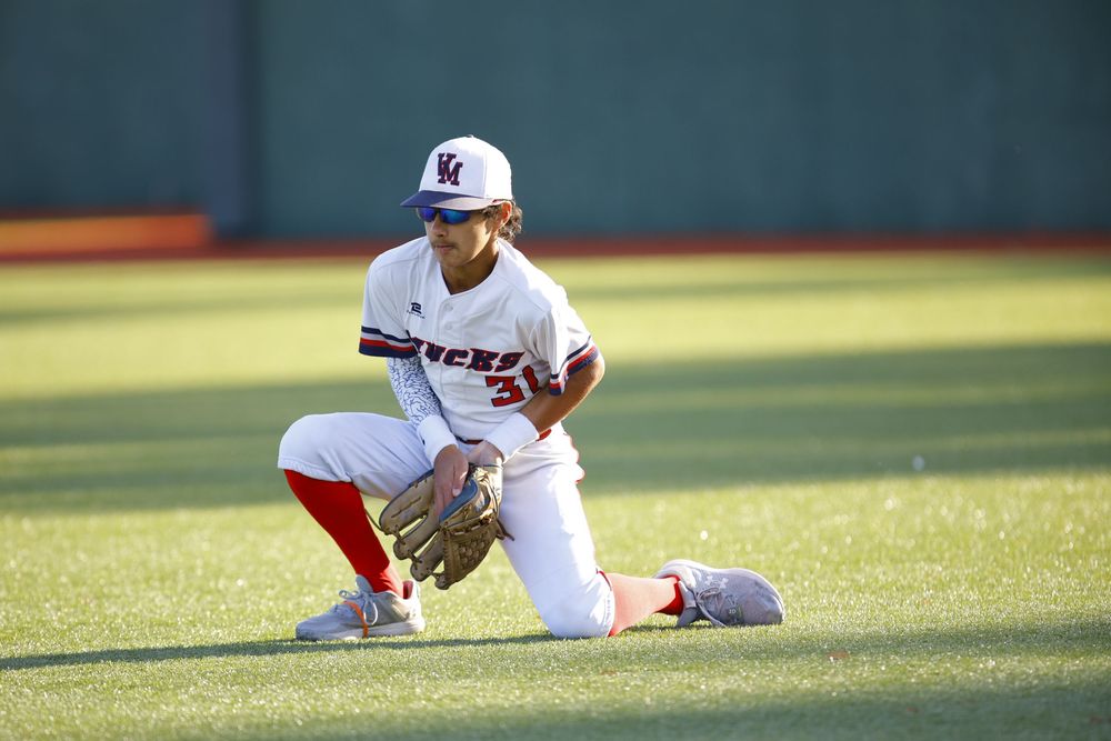 Baseball player in white uniform crouches in fielding position on sunny field.