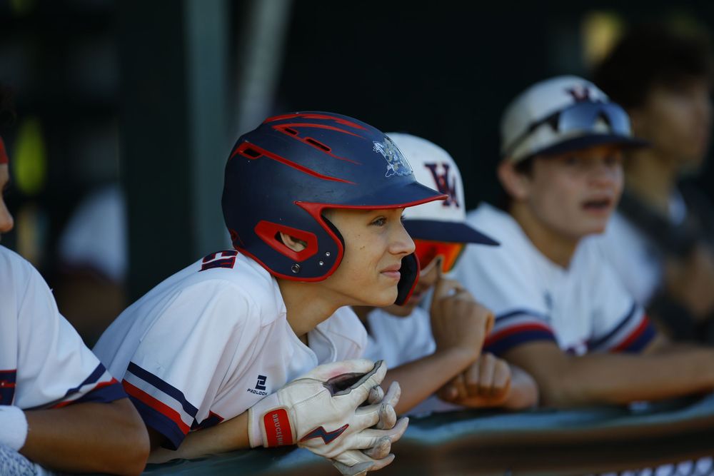 Youth baseball players in white uniforms and navy helmets watch intently from the dugout.