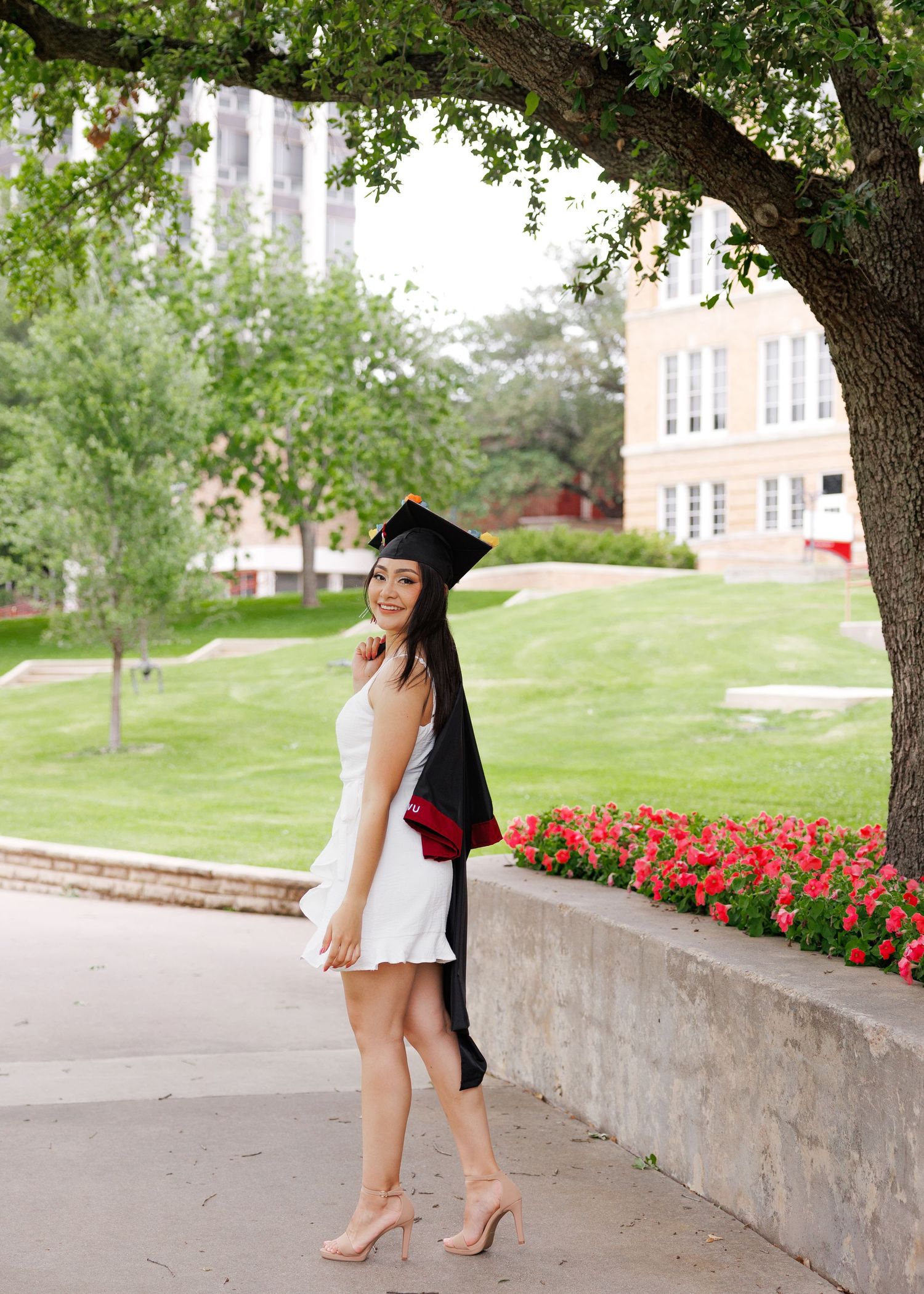 Student in white dress and graduation cap poses on campus walkway with stone wall and red flowers in background.