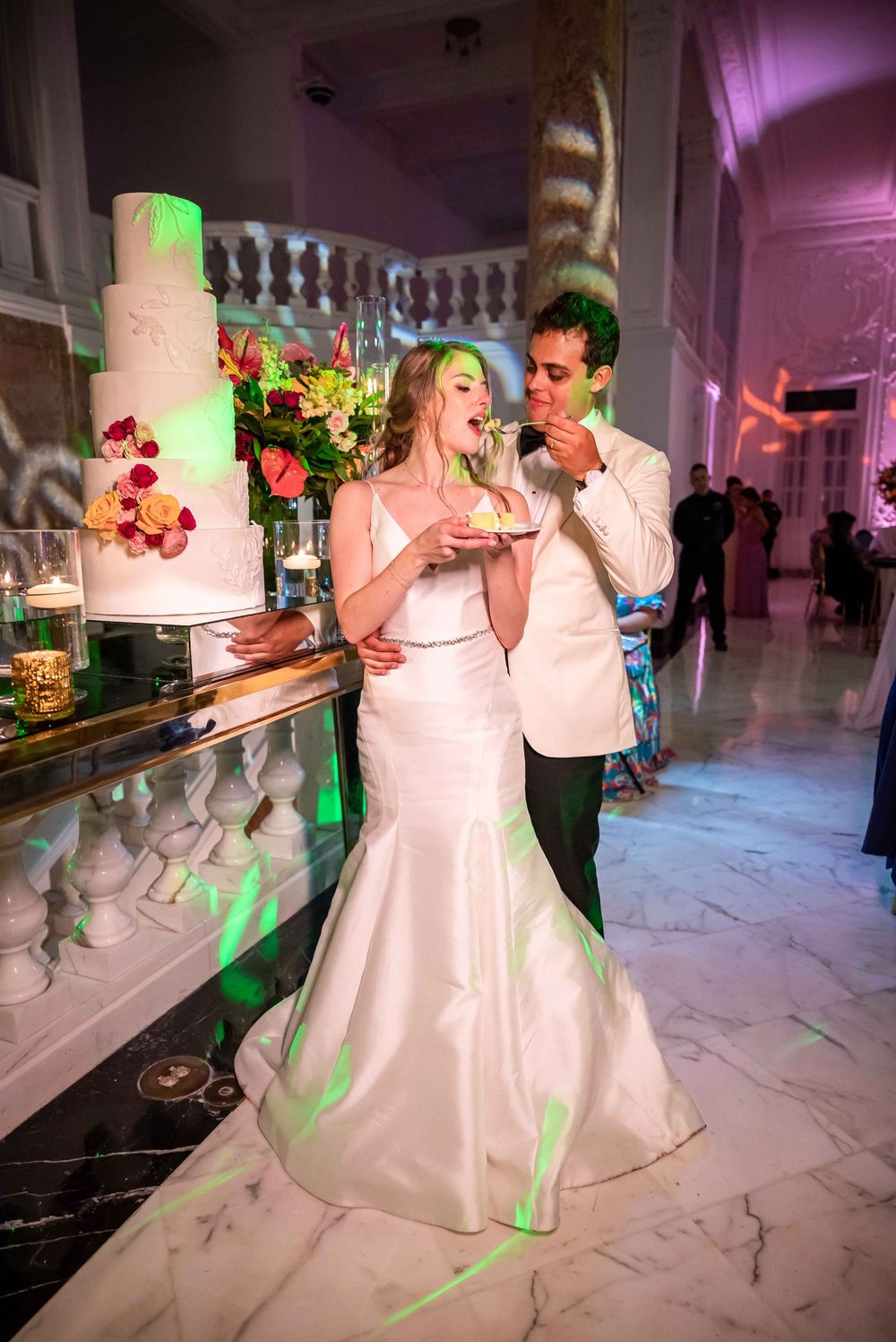 A couple shares a bite of wedding cake together at their reception in an elegant marble venue with purple lighting.