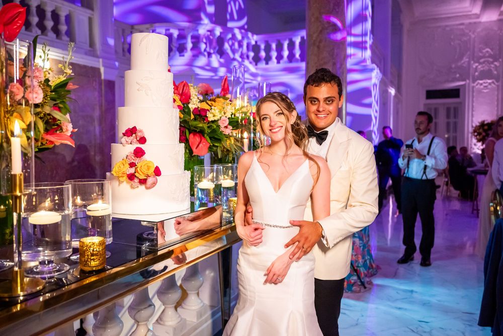 Elegant wedding reception with white tiered cake and bar setup illuminated by purple lighting in ornate venue.