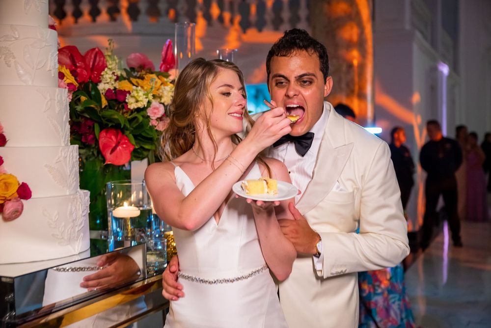 A newlywed couple shares their wedding cake during the reception celebration with floral decorations in the background.
