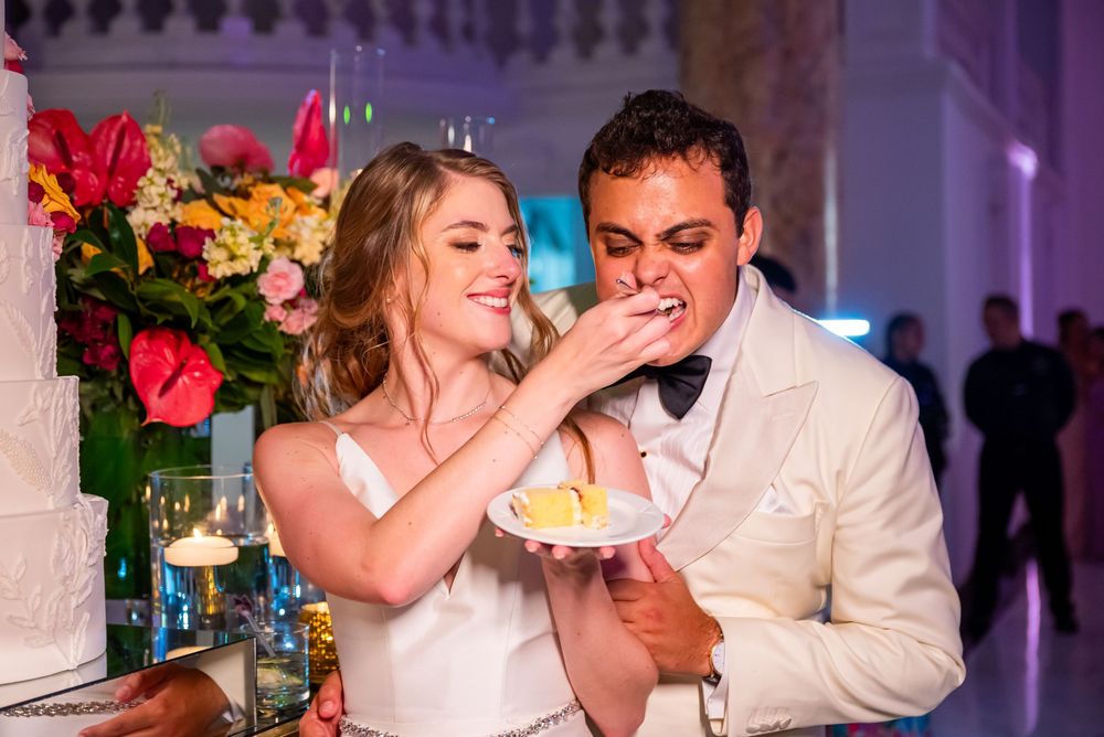 A couple in white wedding attire share a slice of cake at their wedding reception with flowers in the background.