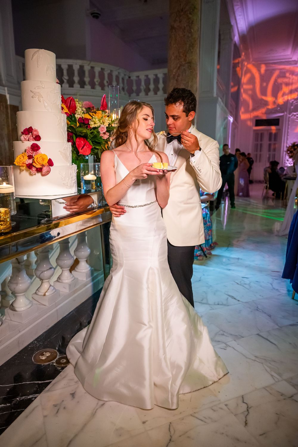 A couple in wedding attire share an intimate moment while drinking wine at an elegant reception with marble floors and decor.