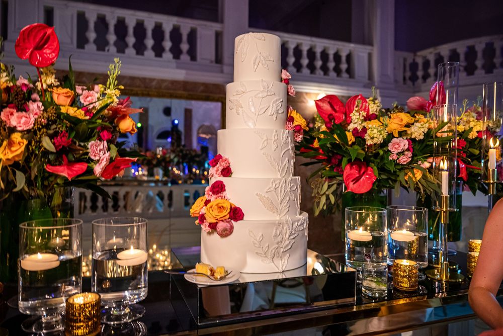 An elegant white wedding cake surrounded by vibrant floral arrangements and glowing candles on a mirrored table at a reception.