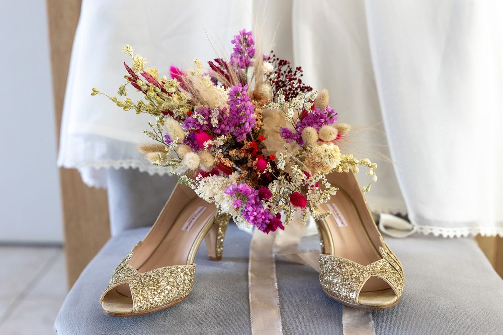 Gold glitter wedding shoes with purple and cream floral bouquet arranged on white surface.