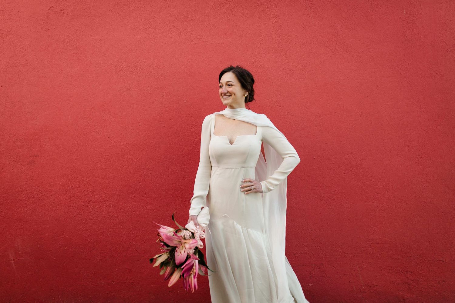 Bride in long-sleeved white wedding dress holds pink bouquet while posing against vibrant red wall.