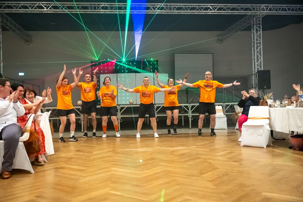 A group in matching orange shirts performs a synchronized dance move at an indoor celebration event.