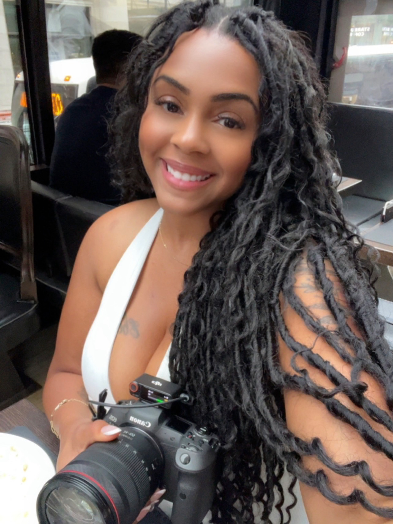 Smiling woman with long locs holding a Canon DSLR camera at a restaurant.