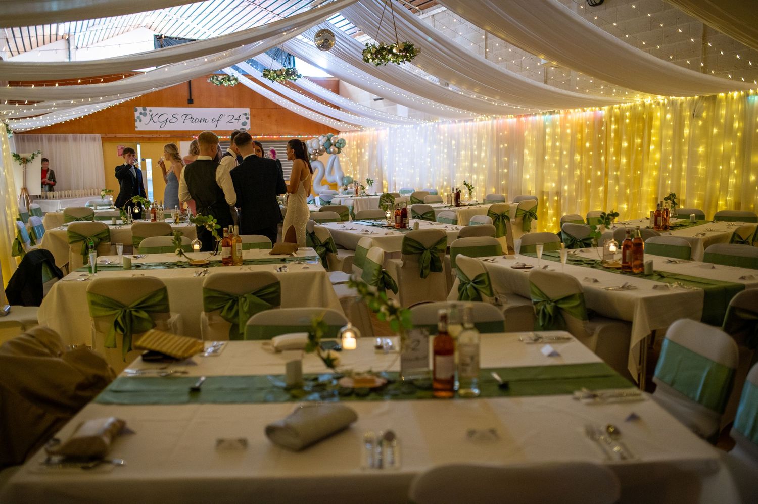 A decorated wedding reception venue with white draped ceiling, string lights, and elegant table settings in sage green.
