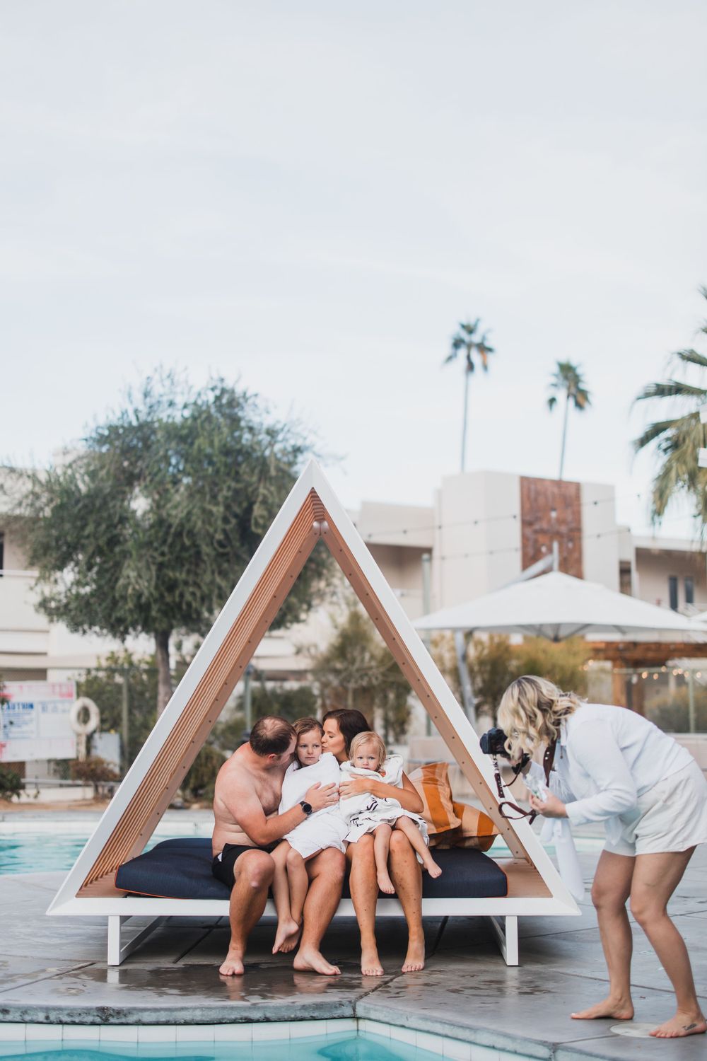 A poolside scene with people relaxing on a triangular lounge bed near palm trees.
