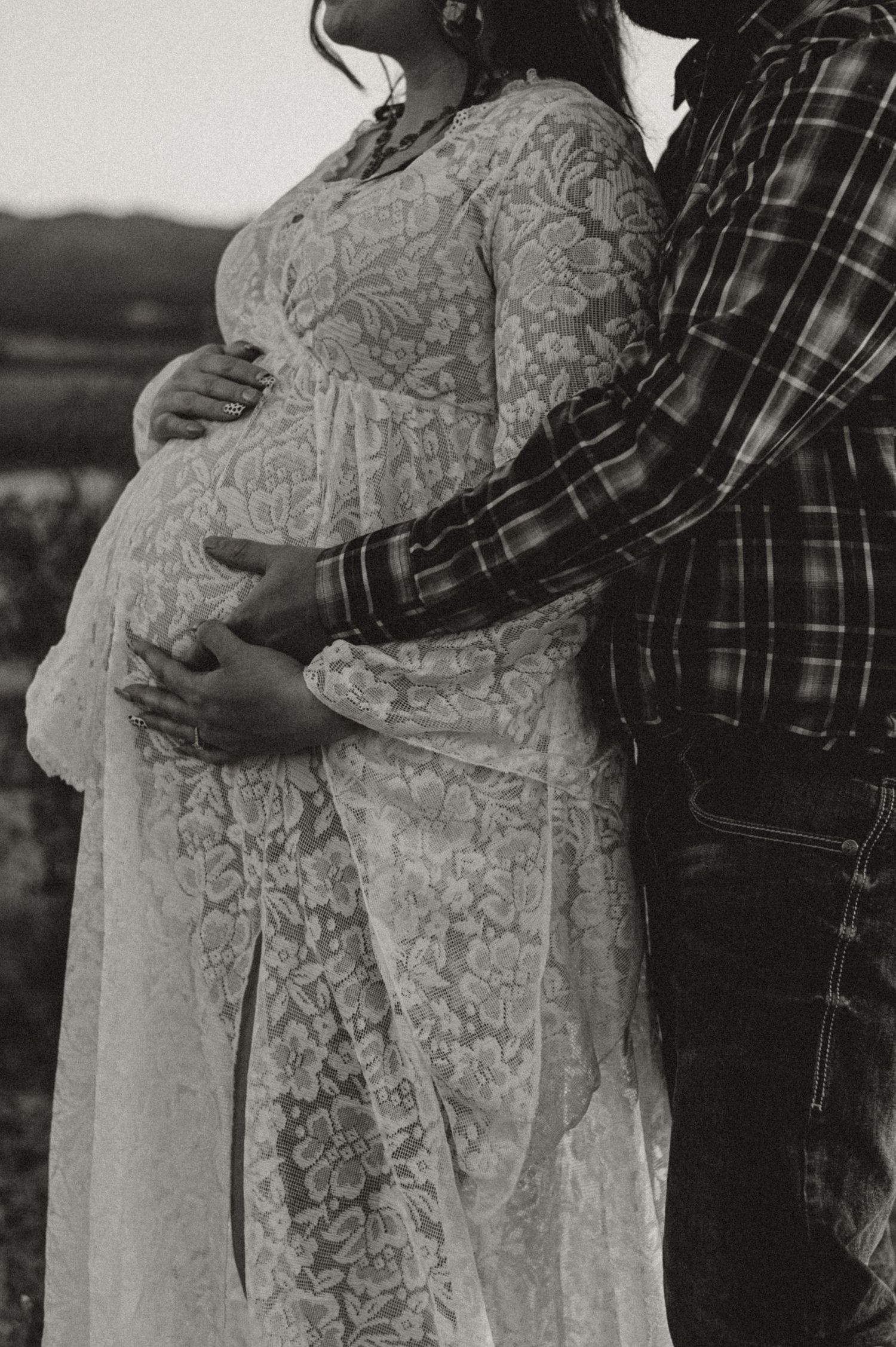 A black and white maternity photo showing a couple's embrace in a lace dress against mountain backdrop.