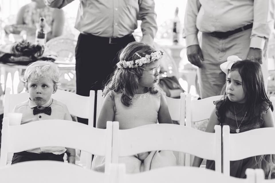 Three children sit in white chairs at what appears to be a formal event, with a child in the middle wearing a floral crown.