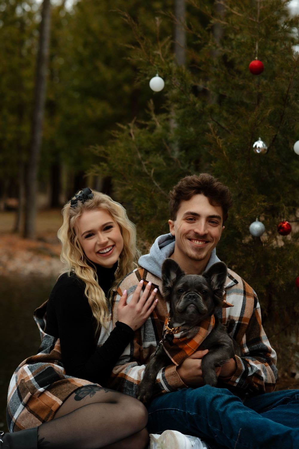 Couple with French Bulldog poses in front of Christmas tree outdoors wearing coordinating plaid outfits.