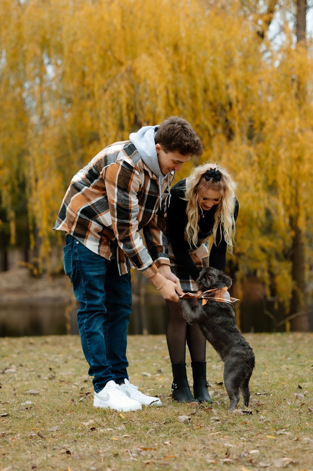 A couple plays with their black French Bulldog puppy under golden autumn willow trees near a pond.
