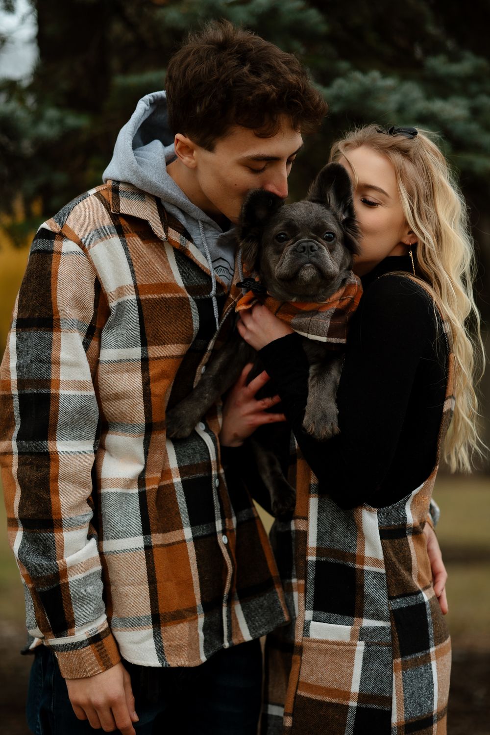 Couple in matching plaid outfits cuddles with their French Bulldog in a park.
