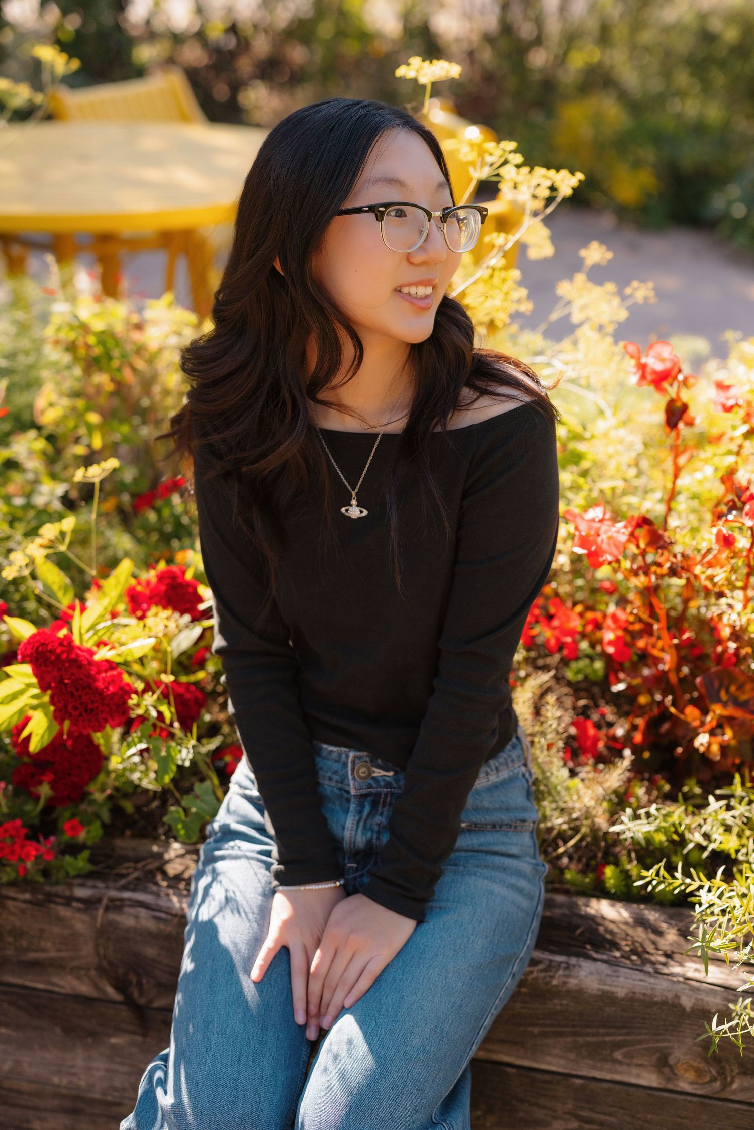 Woman in casual attire smiles while sitting on wooden ledge surrounded by flowers.