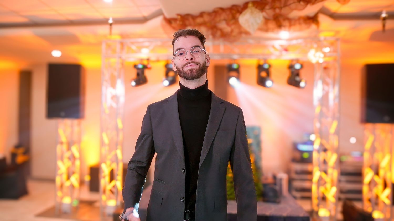 Someone in a grey blazer and black turtleneck stands in an event space with warm lighting and decorative elements.